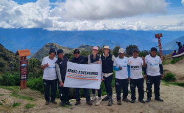 A group of tourists holding a Kenko Adventures flag in the Sacred Valley of the Incas, with terraced hills and towering mountains in the background.